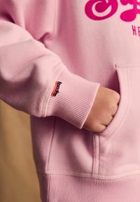 Ružová mikina so klokanie kapsou, rebrovanými manžetami a výrazným logom „Superdry“ v žiarivej ružovej farbe. Mikina má mäkkú textúru.
