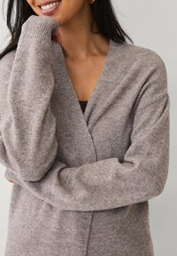 Light brown cardigan made of soft knit material. Features a V-neck, dropped shoulders, and ribbed cuffs. Subtle texture with a cozy appearance.
