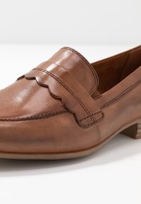 Mocassins en cuir marron avec des détails festonnés, un talon bas empilé, une texture lisse et des coutures sur les côtés. Design classique, style décontracté.