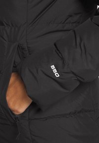 Main d'une personne à l'intérieur de la poche zippée d'une veste matelassée noire avec "550" brodé sur la manche.