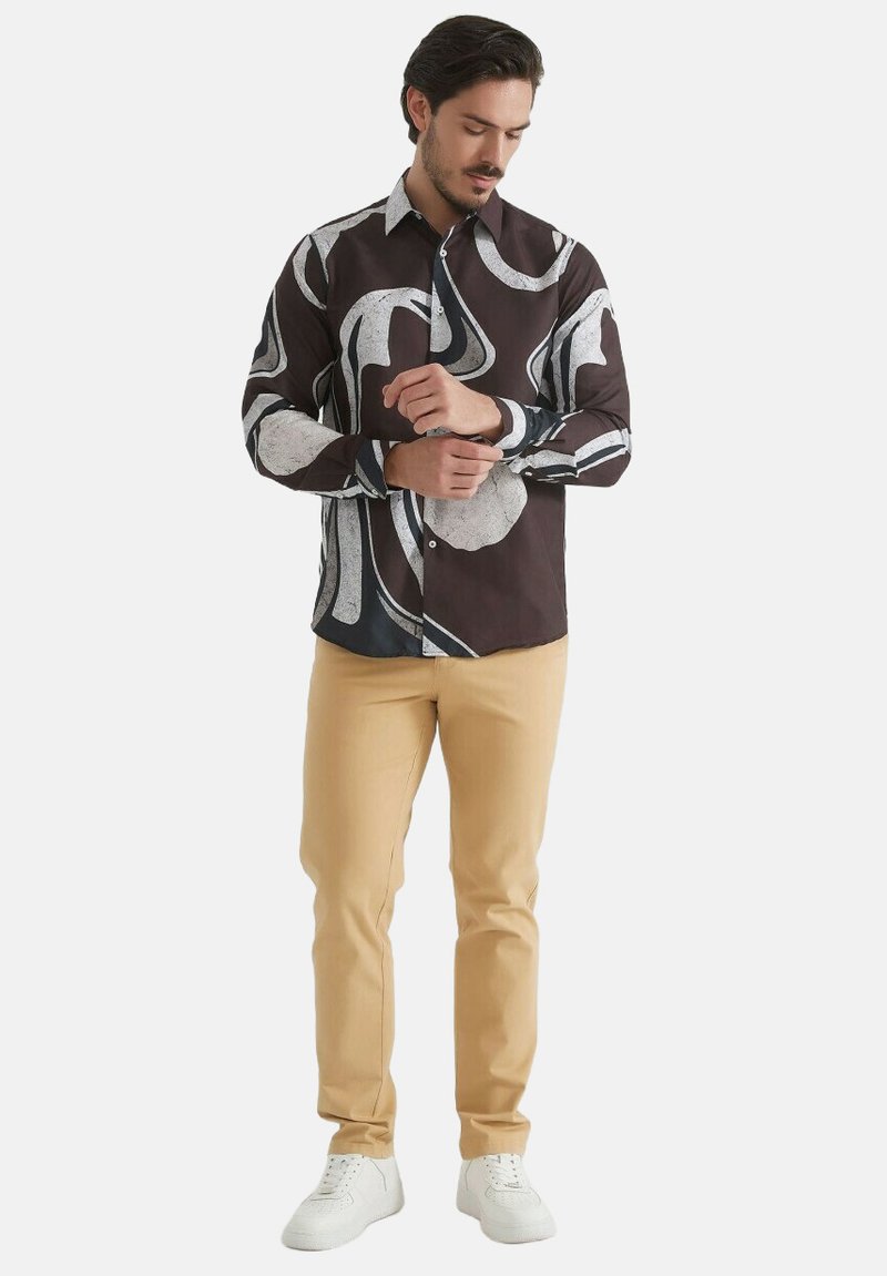 Chemise à manches longues en tissu marron avec des motifs abstraits blancs, associée à un pantalon beige et des baskets blanches.