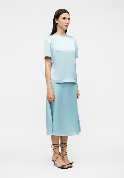 Γυναίκα που φοράει ανοιχτό μπλε σατέν τοπ με κοντά μανίκια και αντίστοιχη μίντι φούστα, σε συνδυασμό με μαύρα σανδάλια με λουράκια και τακούνια, στέκεται μπροστά σε λευκό φόντο.