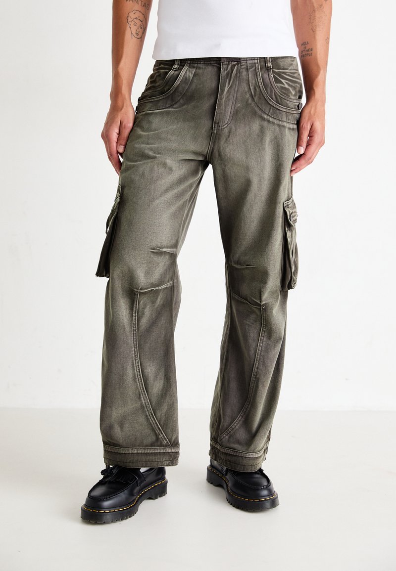 Pantalons cargo en denim vert olive délavé avec plusieurs poches utilitaires, des détails de couture distinctifs et une coupe décontractée. Les chaussures sont en cuir noir.