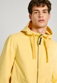Žltá bundu na zips s kapucňou, obsahujúca šnúru na stiahnutie a lesklý zips. Vyrobená z ľahkého materiálu s hladkou textura.
