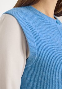 Gros plan sur une personne portant un gilet sans manches en tricot côtelé bleu clair par-dessus une chemise beige à manches longues, montrant les détails de l'épaule et de l'encolure.
