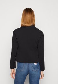 Blazer nero accorciato con colletto a scendere, maniche lunghe, vestibilità strutturata e texture liscia. Abbinato a jeans blu a vita alta.