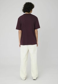 T-shirt en coton bordeaux avec un col rond et des manches courtes, associé à un pantalon blanc ample. L'arrière-plan est gris uni.