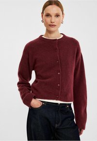 Burgundy kort cardigan med rund hals, långa ärmar och sex framskjutna knappar; mjukt, strukturerat stickat tyg. Bärs över mörka jeans.