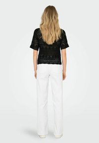 Žena s dlouhými blond vlasy, oblečená v černém krátkém crochtovém topu a bílých kalhotách s vysokým pasem a širokými nohavicemi, stojí otočená zády.