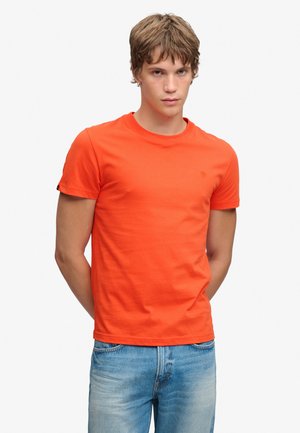 Młody mężczyzna o krótkich brązowych włosach, ubrany w pomarańczowy t-shirt z krótkim rękawem i niebieskie dżinsy, stoi z rękami za plecami.