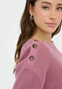 Femme portant un pull en maille rose texturé avec trois boutons en écaille de tortue sur l'épaule, des boucles d'oreilles créoles en or et un collier chaîne en or.