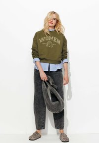 Sweatshirt vert olive avec "FEMMES" imprimé sur le devant, superposé sur une chemise bleu clair à boutons, accompagné d'un jean en denim foncé et de chaussures à imprimé léopard.