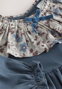 Modré šaty s květinovým, volánkovým vrškem z lehké šedé látky s modrými květinovými vzory; doplněné modrým sametovým mašle.