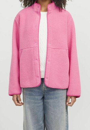 Personne portant une veste rose vif texturée à boutons-pression par-dessus un pull en maille torsadée blanc et un jean bleu clair, debout devant un fond uni.