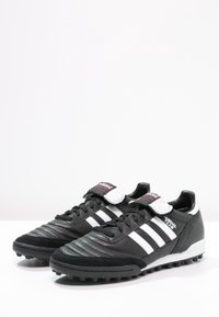 Chaussures de football Adidas Mundial Team noires avec bandes blanches et fermeture Velcro, conçues pour jouer sur gazon synthétique.