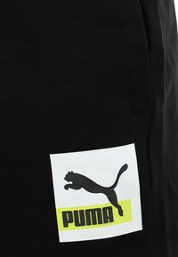 Pantaloni corti neri con un grande logo quadrato bianco che presenta un gatto nero che salta e la parola "PUMA" in lettere in grassetto nere e gialle.