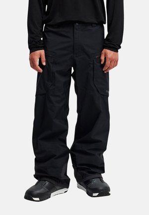 Homme portant un pantalon cargo noir avec des poches zippées et des chaussures noires, debout devant un fond clair uni.