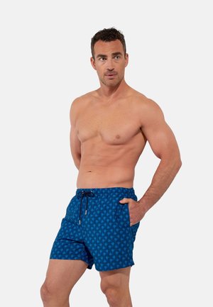 Mężczyzna bez koszulki, o krótkich ciemnych włosach, ubrany w niebieskie kąpielówki z wzorem, stoi z jedną ręką w kieszeni i patrzy w bok.