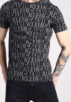 T-shirt Calvin Klein noir et gris coupe slim avec le nom de la marque en motif répété porté par une personne ayant l'avant-bras droit tatoué et un jean foncé.