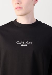 Crna pamučna majica s okruglim izrezom, s bijelim natpisom "Calvin Klein JEANS" otisnutim na prsima. Glatka tekstura, minimalistički dizajn.