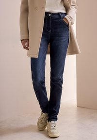 Mörkblå denimjeans med smal passform, med en framficka, knappstängning och kontrasterande sömmar, tillsammans med beige sneakers.