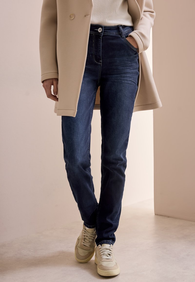 Mörkblå denimjeans med smal passform, med en framficka, knappstängning och kontrasterande sömmar, tillsammans med beige sneakers.