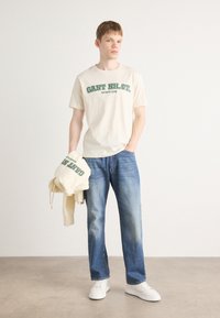 Beige cotton t-shirt with green lettering, paired with light blue denim jeans. A beige hoodie is held in one hand. White sneakers complete the outfit.