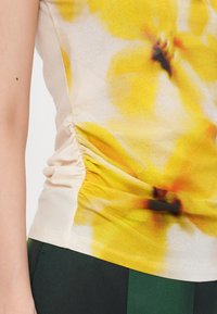 Tett gul blomstret topp med samlet sidedeatil, laget av mykt stoff. Har en jevn tekstur og livlige, blandede gule og oransje mønstre.
