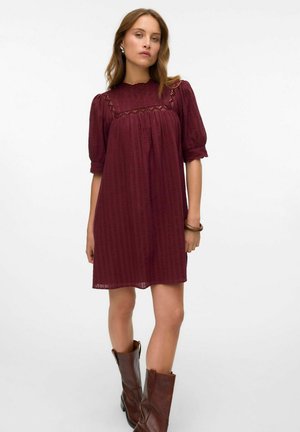 Vestito bordeaux a maniche puff con un accento in pizzo sul colletto, realizzato in tessuto testurizzato, indossato con stivali al ginocchio in pelle marrone.