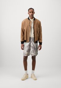 Brown suede bomber jacket, cream turtleneck, camo-patterned shorts, and beige sneakers with textured details. Matching white socks visible.