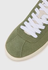 Sapatilhas verdes em camurça com atacadores brancos, bico arredondado, sola contrastante na cor creme e detalhes de costura subtil ao longo do design.