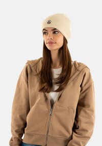 Giovane donna con lunghi capelli castani, che indossa un berretto beige e una felpa con zip marrone chiaro, che guarda leggermente di lato su uno sfondo bianco.