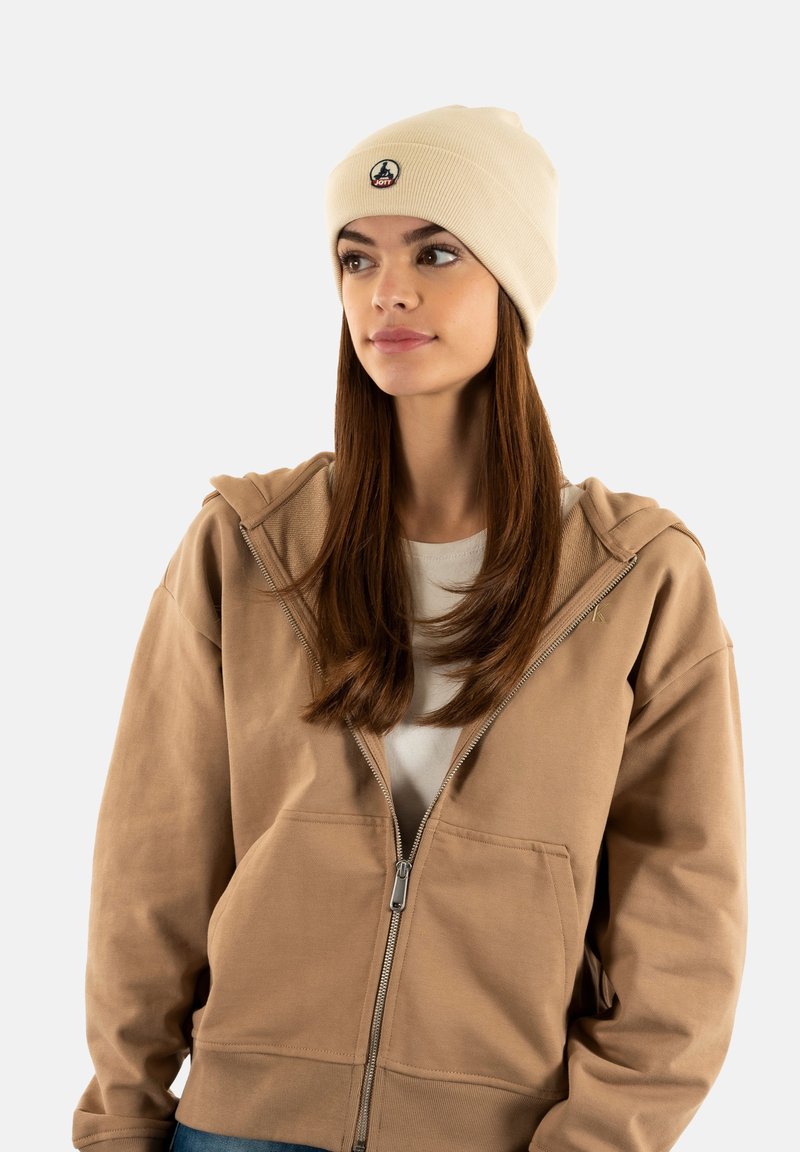 Mujer joven con cabello largo castaño, llevando un gorro beige y una sudadera con cremallera de color marrón claro, mirando ligeramente hacia un lado contra un fondo blanco.