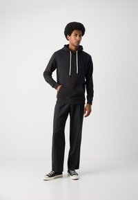 Haut à capuche noir avec une poche kangourou et des cordons de serrage blancs, associé à un pantalon sombre ajusté et des baskets noires avec des semelles blanches.