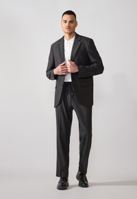 Blazer gris ajustado sobre una camiseta polo blanca y pantalones grises con cordón. Zapatos negros completan el conjunto. Estilo minimalista, texturas suaves.