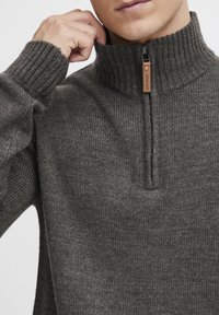 Pull-over en tricot gris avec un col montant et une fermeture éclair quart. Présente des poignets côtelés et un tissu texturé. Accentué d'une languette en cuir.