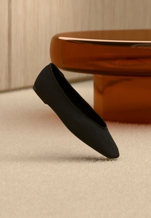 Chaussure plate noire à bout pointu sur un tapis beige, avec une table basse ronde translucide de couleur ambre en arrière-plan.