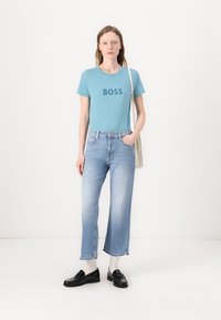 Šviesiai mėlyna marškinėlių su užrašu „BOSS“, derinami su išblukusiais mėlynais platėjančiais džinsais ir juodais loafers. Modelis laiko kreminį rankinį maišą.