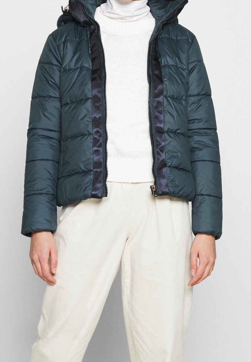 Tealfärgad puffjacka med dragkedja fram, huva och ribbade ärmslut med horisontell söm. Lagd över en vit tröja.