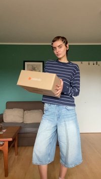 Pappkarton mit Zalando-Logo, gehalten von einer Person in einem navy-blauen gestreiften Langarmhemd und hellblauen Jeansculottes, die drinnen steht.