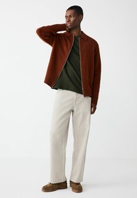 Rust-colored knitted zip cardigan with a collar, worn over a dark green shirt and light beige pleated trousers, paired with brown shoes.