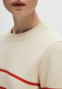 Cremefarbener gerippter Pullover mit rundem Ausschnitt und zwei horizontalen roten Streifen über der Brust, der eine glatte Textur aufweist.