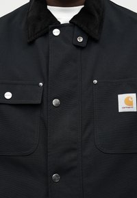 Μαύρο μπουφάν Carhartt με κουμπιά κούμπωμα, δύο μπροστινές τσέπες και γιακά από κοτλέ, φορεμένο πάνω από ένα λευκό πουκάμισο.