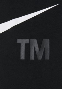 Vit Nike-svans över grå "TM"-bokstäver på svart texturerad tygbakgrund.