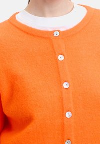 Nära bild av en person som bär en orange knäppt kofta över en vit rundhalsad tröja, som visar övre delen av bröstet och nackområdet.