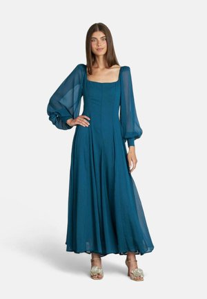 Abito lungo in teal con maniche a sbuffo trasparenti, scollo a quadrato e gonna plissettata. Il materiale sembra leggero e fluido.