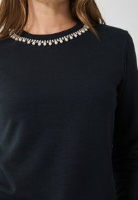 Navy kék hosszú ujjú felső, kerek nyakkivágással, amelyet díszített gyöngyök és gyöngyök sora ékesít, kiemelve egy elegáns részletet.