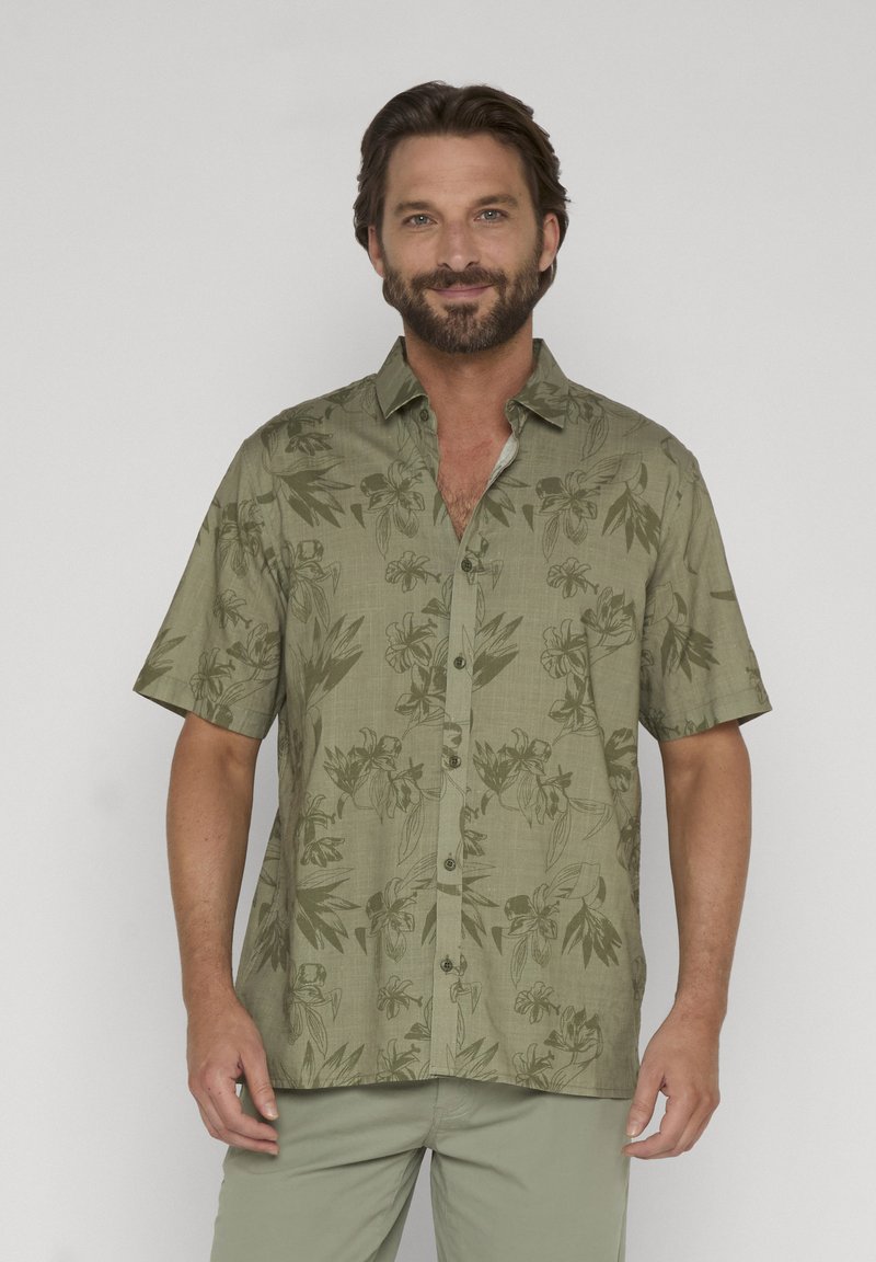 Homme barbu portant une chemise verte à manches courtes avec un motif floral, debout devant un fond clair uni.