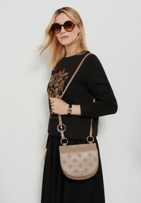 Beżowa skórzana torebka na ramię z wytłoczonym kwiatowym wzorem, okrągłym kształtem i metalowymi akcentami. Przypinany regulowany pasek.