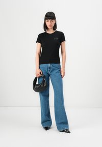 Giovane donna in piedi con una t-shirt nera GAP, jeans blu larghi, scarpe nere con punta, mentre tiene una piccola borsa in pelle nera su uno sfondo neutro.